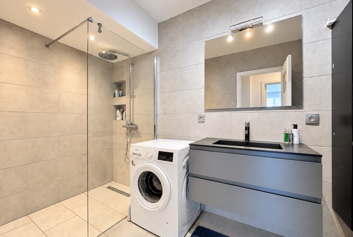 Modern bathroom with glass shower enclosure to the left, rainfall showerhead, and built-in shelves; washing machine beside a gray vanity and large mirror.