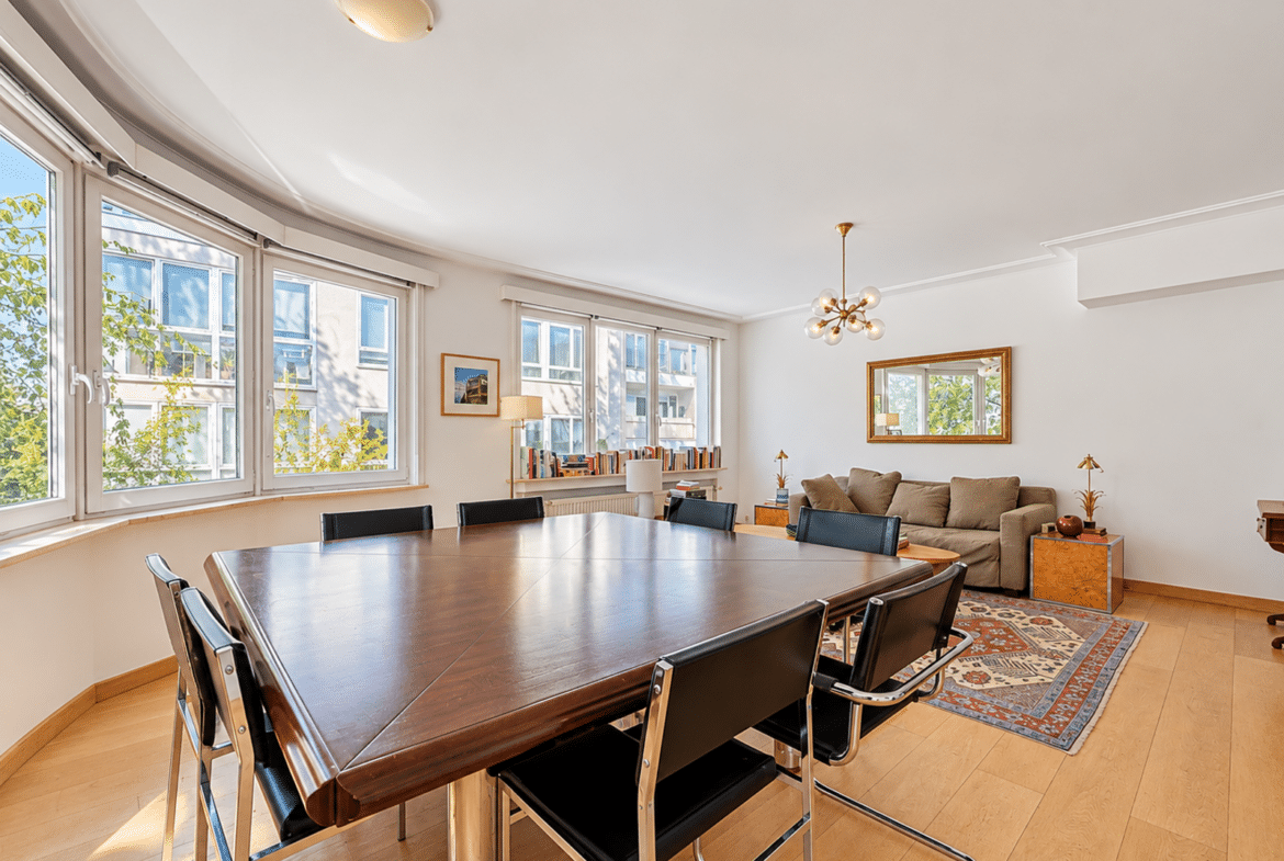 Bright dining room with a long wooden table and black leather chairs, curved bay windows, and a seating area with a sofa.