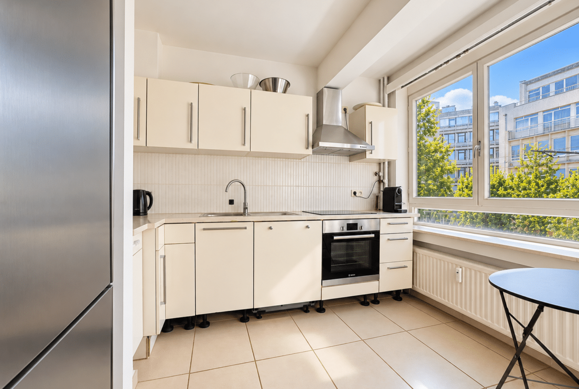 Bright, modern white kitchen with stainless steel appliances, a large window view of trees and buildings, and a small round table by the radiator