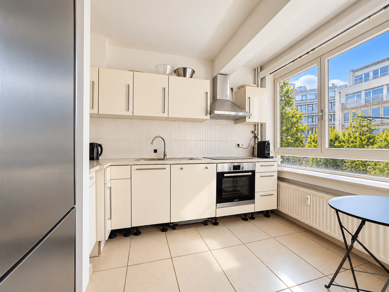 whise photo 7 Bright, modern white kitchen with stainless steel appliances, a large window view of trees and buildings, and a small round table by the radiator