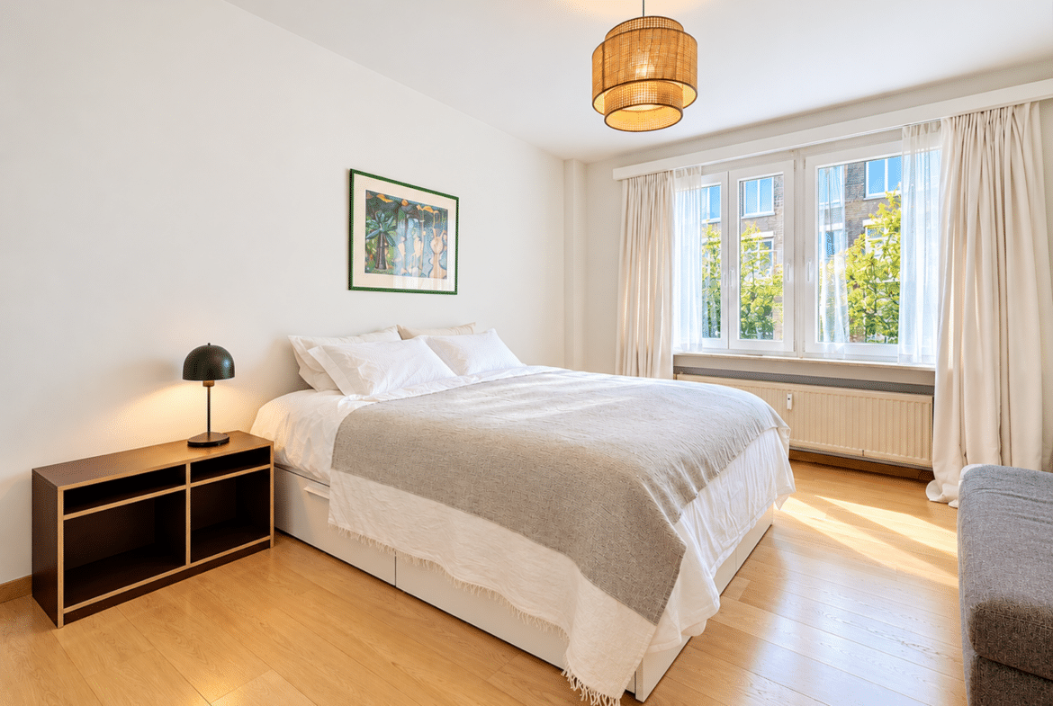 Bright, modern bedroom with a large white bed, white linens, and a wooden nightstand with a black lamp beside a framed tropical artwork on the wall. A large window with sheer curtains fills the room with natural light.