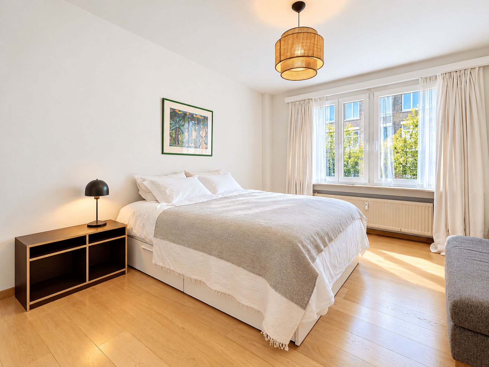 whise photo 8 Bright, modern bedroom with a large white bed, white linens, and a wooden nightstand with a black lamp beside a framed tropical artwork on the wall. A large window with sheer curtains fills the room with natural light.