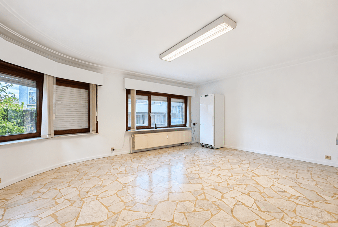 Bright empty room with curved wall, three large brown-framed windows, a radiator beneath, and a tall white cabinet on the right.