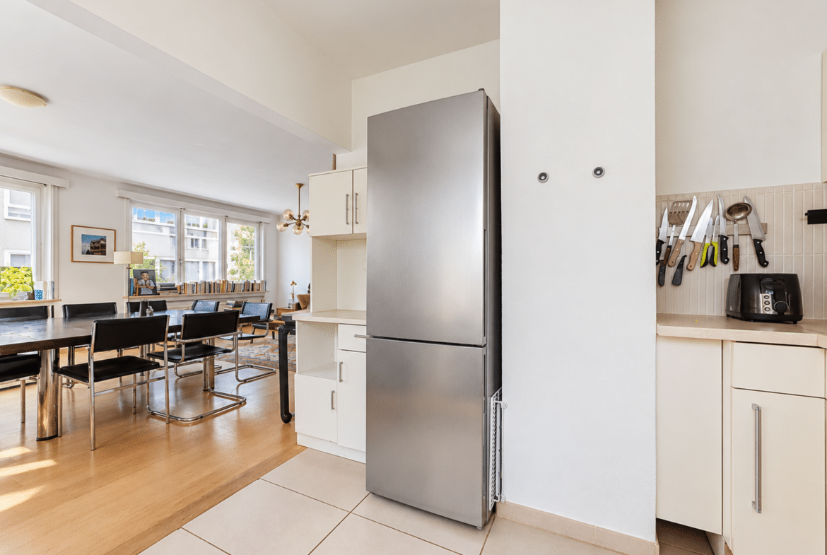 Bright open-plan kitchen and dining area featuring a large stainless steel refrigerator, white cabinets, a wall knife rack, and a long wooden table with black chairs by the windows.