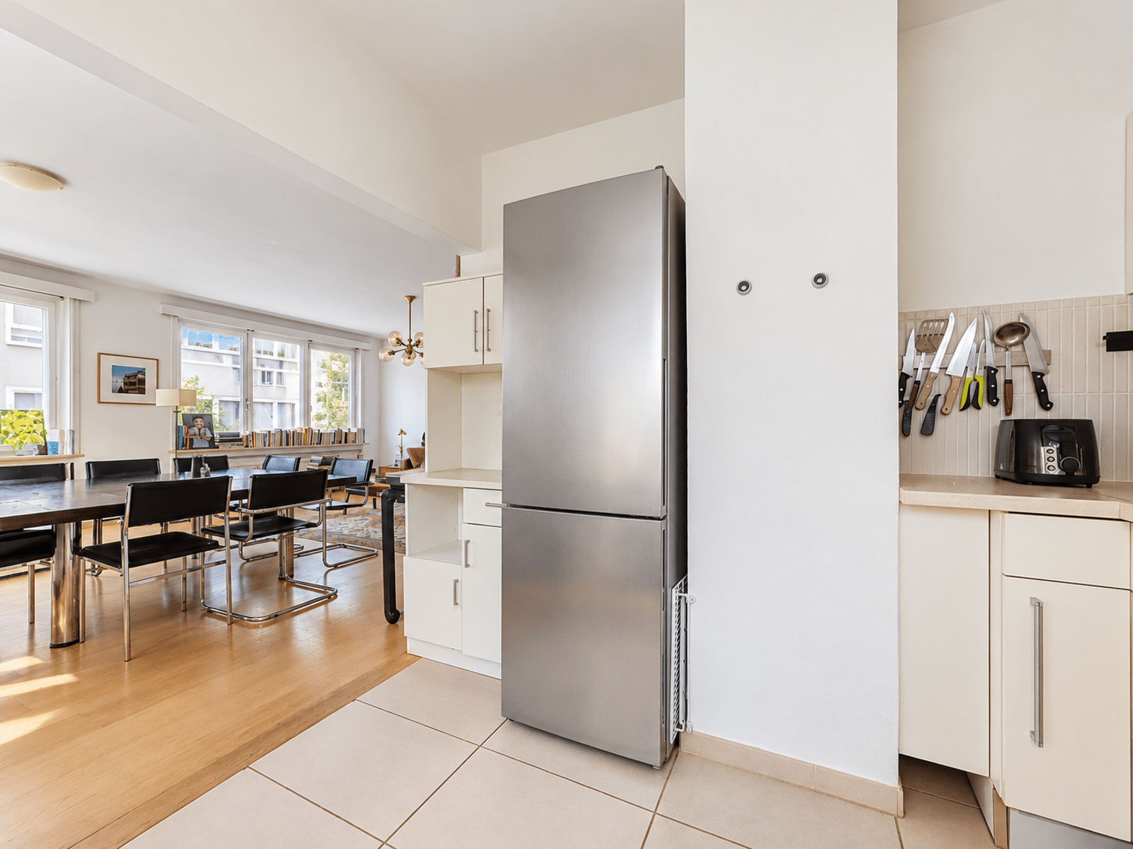 whise photo 6 Bright open-plan kitchen and dining area featuring a large stainless steel refrigerator, white cabinets, a wall knife rack, and a long wooden table with black chairs by the windows.