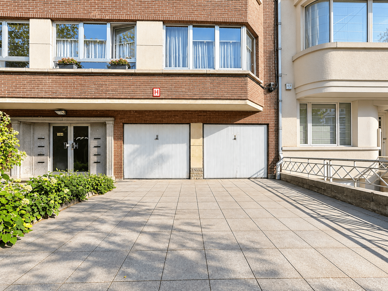 Façade en Briques avec Garages – You Real Estate Agency Façade en briques d’une maison avec garages et entrée pavée.