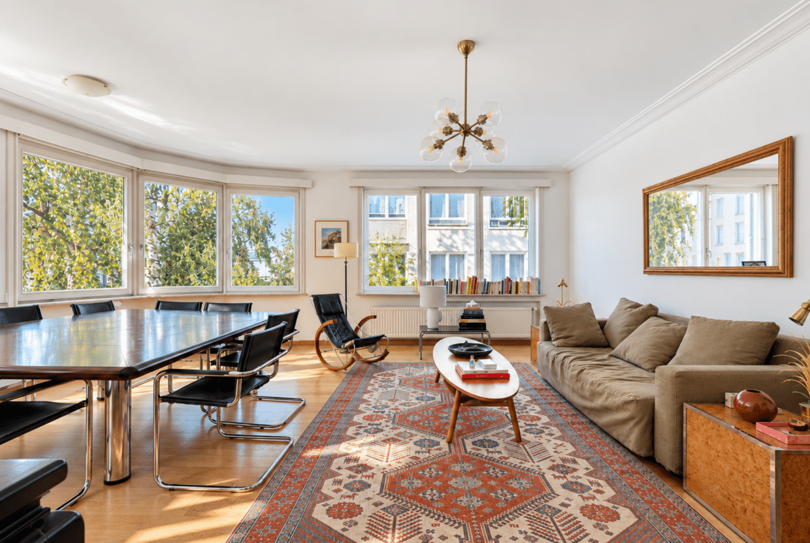 Bright open living and dining room with a long wooden table and black chairs beside a wall of windows, beige sofa, and a patterned rug.