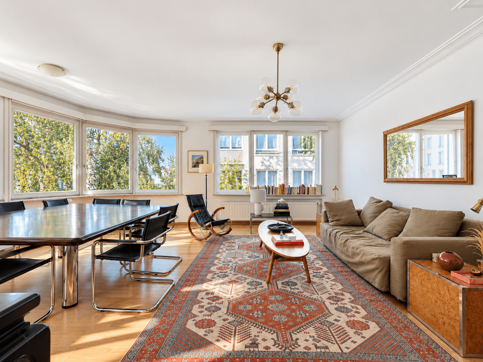 whise photo 2 Bright open living and dining room with a long wooden table and black chairs beside a wall of windows, beige sofa, and a patterned rug.