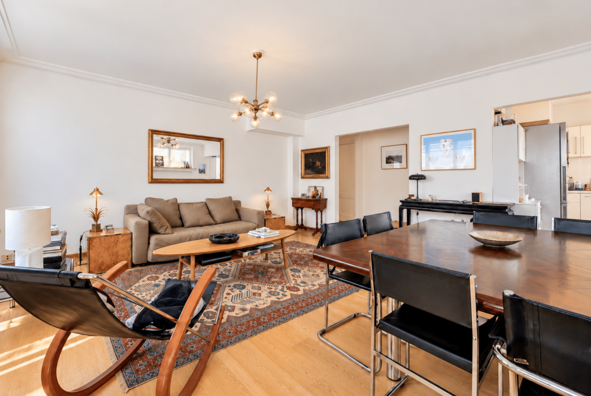 Spacious living and dining room with a beige sofa, wooden coffee table, and a patterned rug under a modern chandelier.