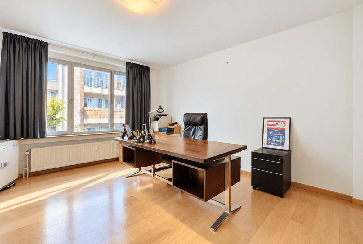 Modern home office with a wooden desk, black leather chair, large window with dark curtains, and a framed poster on a small black cabinet.