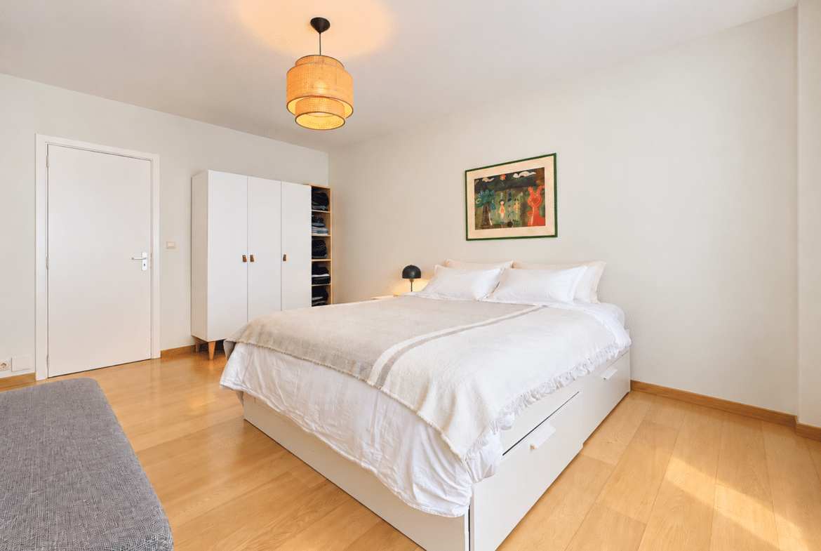 Bright minimalist bedroom with a white bed, white wardrobe, and wooden floor; woven pendant light overhead.