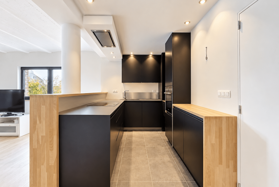 Cuisine moderne en couloir avec mobilier noir et bois, ouverte sur salon lumineux.