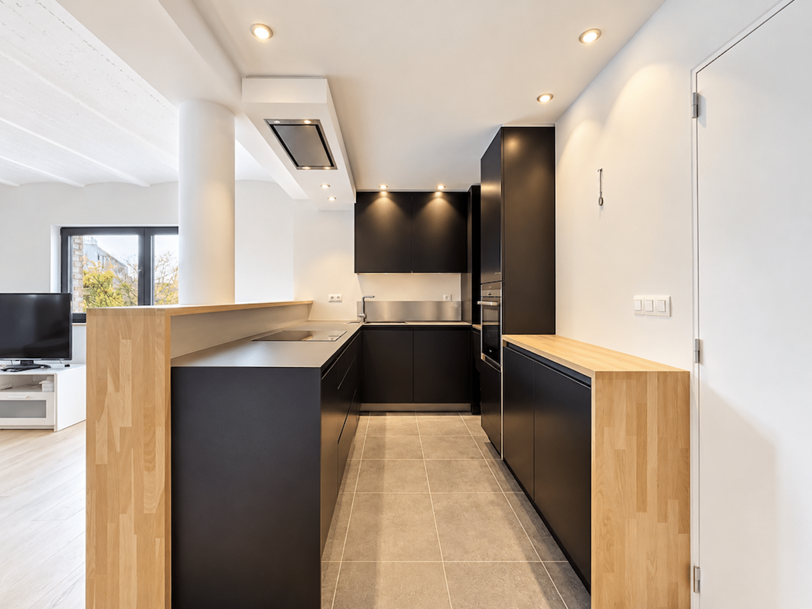 Cuisine Moderne en Couloir – You Real Estate Agency Cuisine moderne en couloir avec mobilier noir et bois, ouverte sur salon lumineux.