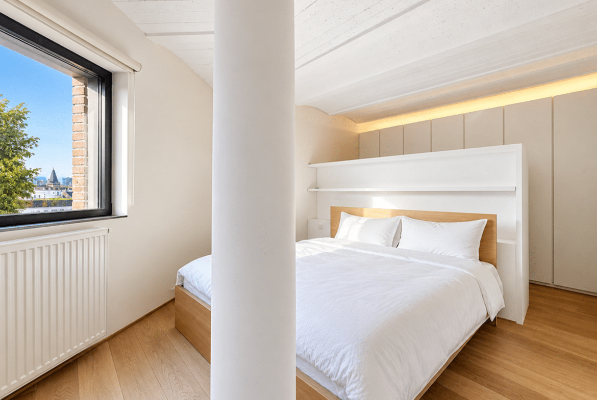 Chambre lumineuse d’appartement moderne avec parquet, lit blanc et grande fenêtre.